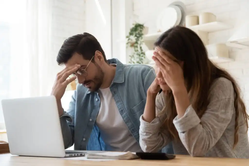 A man and woman sit at a table looking stressed while staring at a laptop. Both hold their heads in their hands, and documents and a calculator are also on the table.