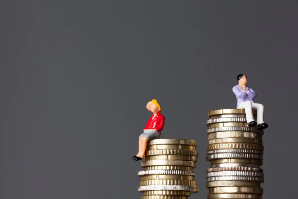 Two small figurines, one of a woman and one of a man, sit separately on top of uneven stacks of coins, with the man on a higher stack, symbolizing gender pay gap. The background is plain gray.
