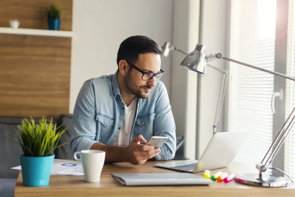A man wearing glasses and a denim shirt sits at a desk with a laptop, holding a smartphone. There’s a coffee mug, a plant, and office supplies on the desk. He looks at the laptop in a bright, modern room.