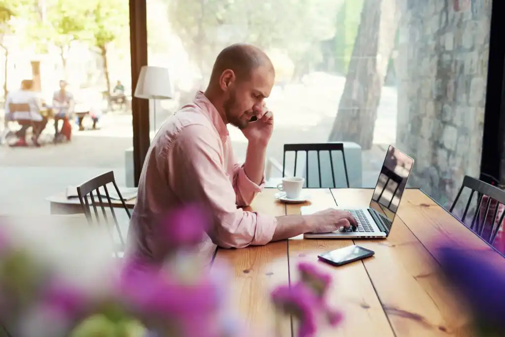 A man sits at a wooden table in a bright café, working on a laptop and holding a phone to his ear. A coffee cup and smartphone are on the table, with pink flowers blurred in the foreground.