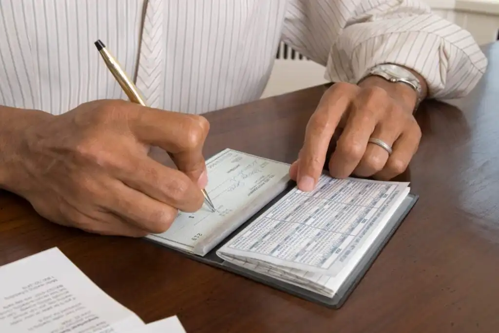 A person wearing a striped shirt and a ring writes a check with a gold pen at a wooden table, with a checkbook and papers nearby.