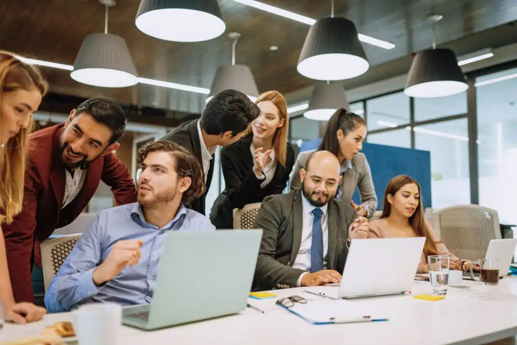 A group of professionals gathers around a table with laptops and documents, engaged in discussion and teamwork in a modern office with hanging lights and wooden ceiling.