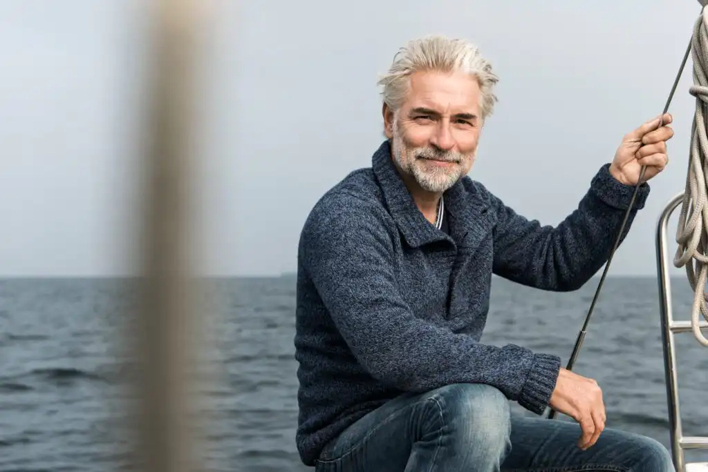 A smiling older man with gray hair and a beard, wearing a blue sweater and jeans, sits on a boat holding a rope with the ocean in the background.