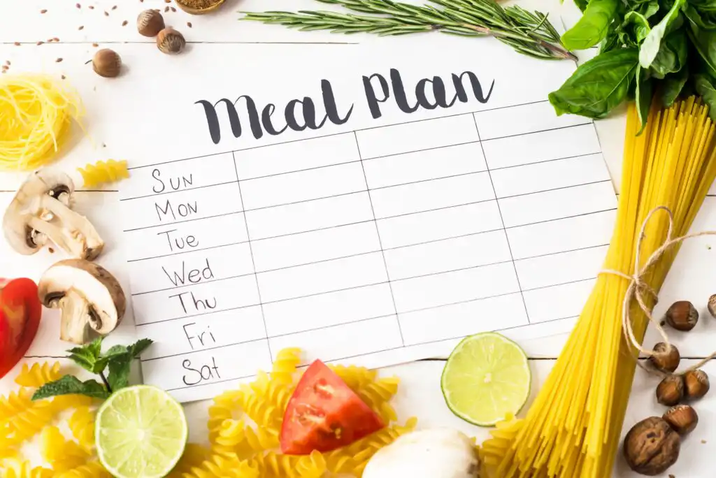 A blank weekly meal plan chart surrounded by fresh ingredients like pasta, tomatoes, mushrooms, lime, basil, and rosemary on a white table. Days of the week are listed along the left side.