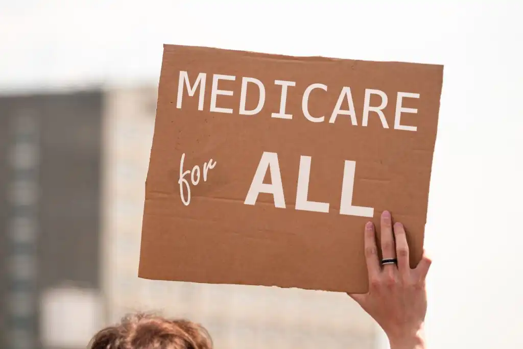 A person holds up a cardboard sign that reads MEDICARE for ALL in large white letters, advocating for universal healthcare.