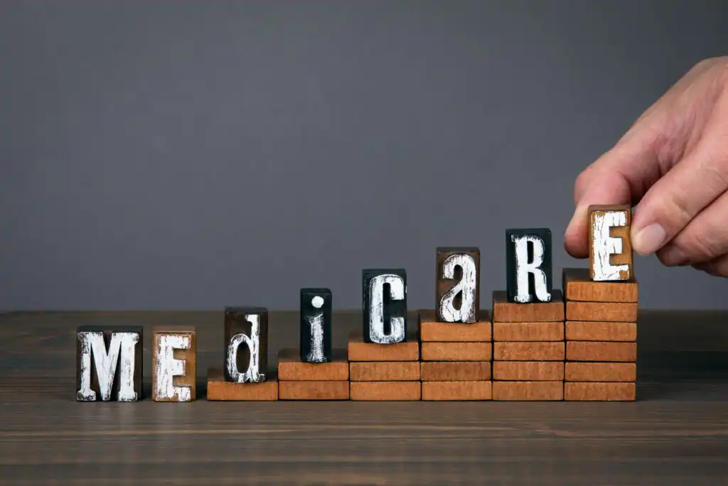 A hand places the last letter block in the word MEDICARE on top of stacked wooden blocks, forming steps rising from left to right against a gray background.