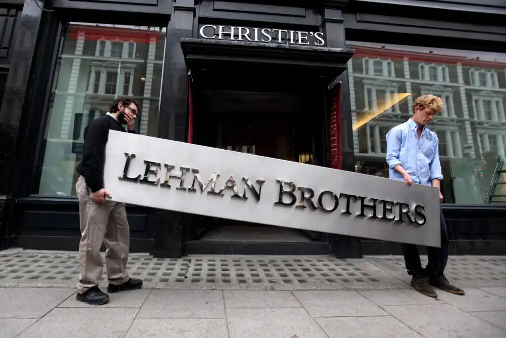 Two men carry a large metallic Lehman Brothers sign outside Christie’s auction house, with the building and its sign visible in the background.