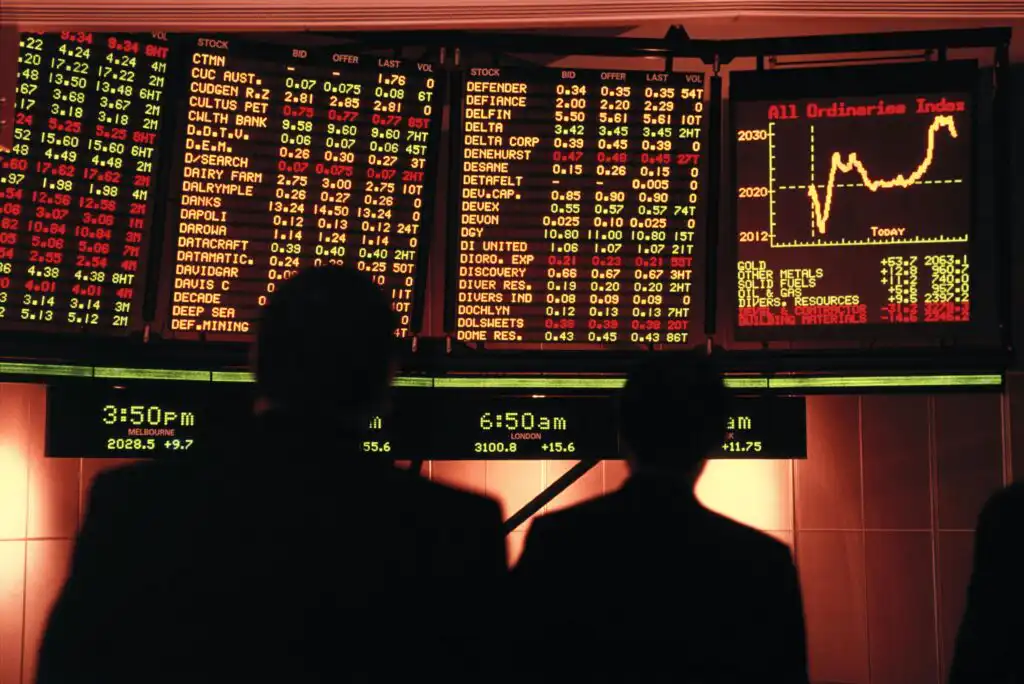Silhouettes of three people stand in front of large, brightly lit electronic stock market boards displaying various stock prices, indices, and a line graph labeled All Ordinaries Index.