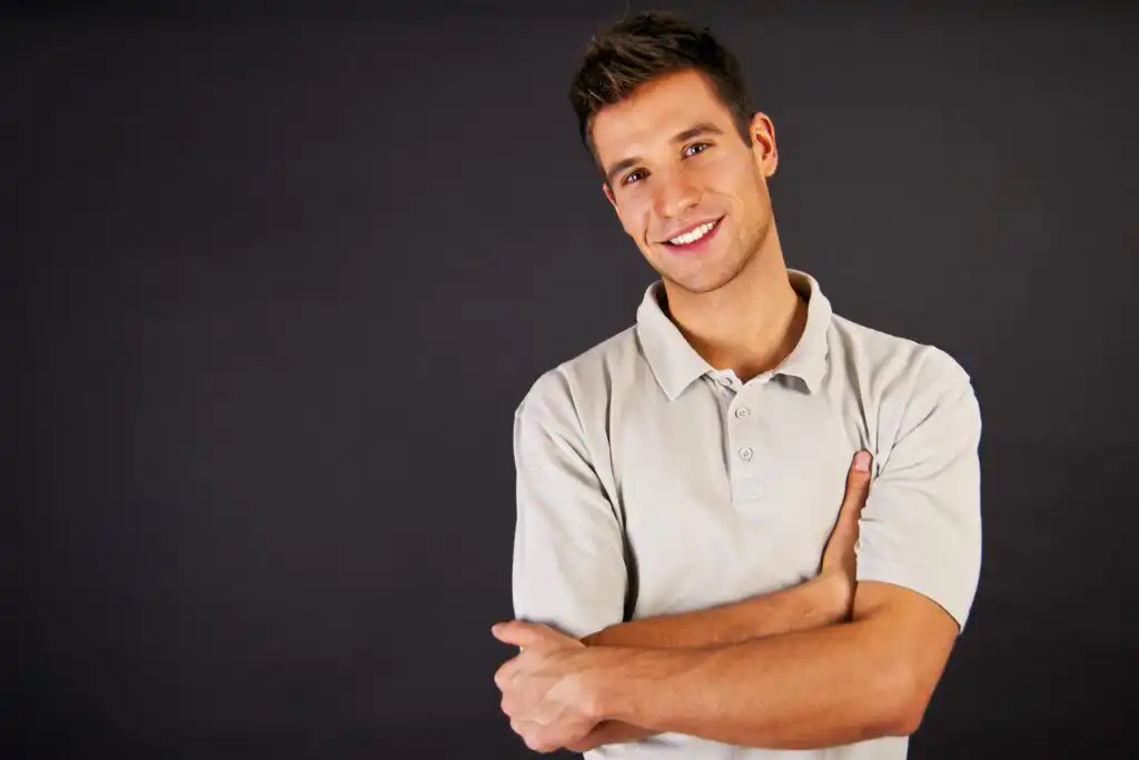 A young man in a light gray polo shirt stands against a plain dark background, smiling with arms crossed and head slightly tilted.