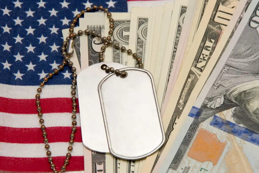 Two blank military dog tags on a chain rest on an American flag beside a spread of assorted US dollar bills.
