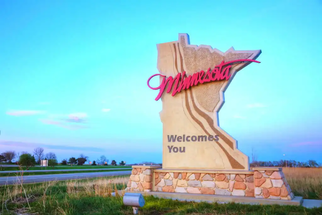 A roadside sign shaped like the state of Minnesota reads Minnesota Welcomes You in large letters, with a blue sky and open fields in the background.