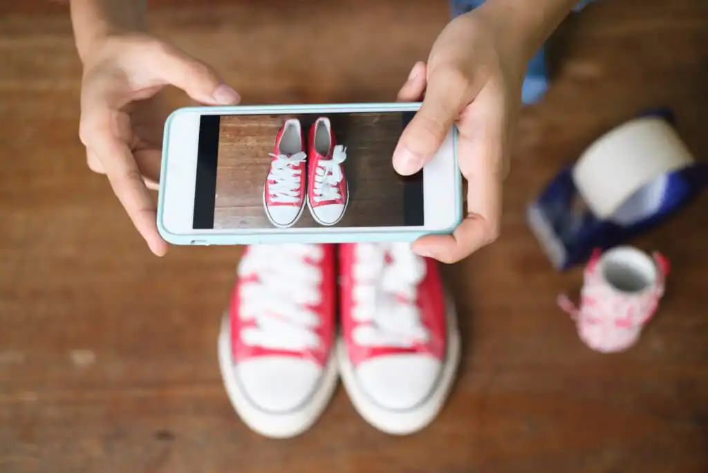 A person holds a smartphone to take a photo of a pair of red and white sneakers on a wooden floor, capturing the shoes directly from above.
