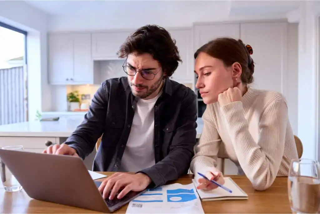 A man and woman sit at a kitchen table, looking at a laptop and documents with charts. The man types on the laptop while the woman holds a pen, appearing thoughtful. Both seem to be concentrating on their work.