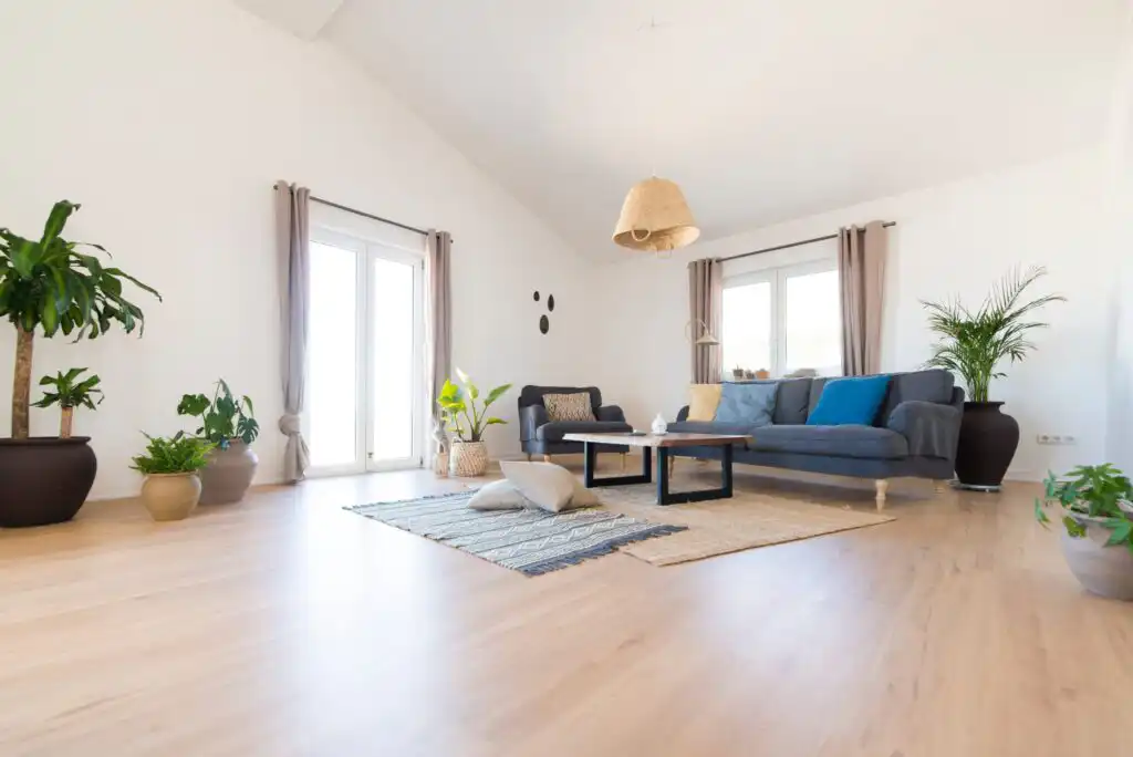 A bright living room with wooden floors, potted plants, a blue-gray sectional sofa with cushions, coffee tables, a woven pendant light, and large windows with sheer curtains letting in natural light.