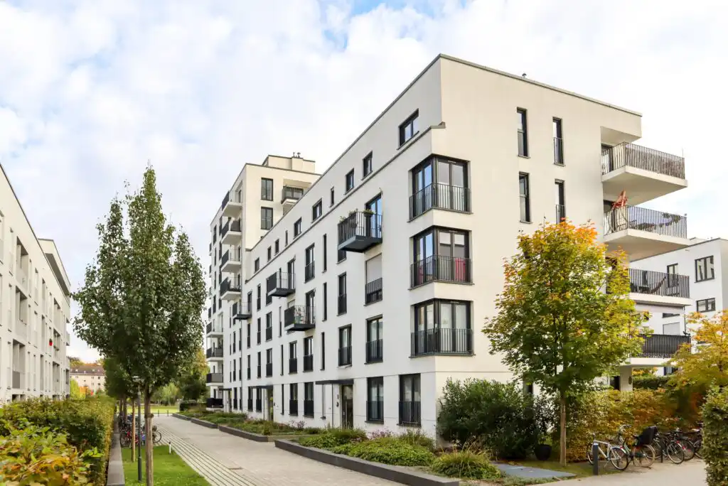 Modern white apartment buildings with multiple balconies, surrounded by greenery and trees, lined along a pedestrian walkway. Bicycles are parked nearby, and the sky is partly cloudy.