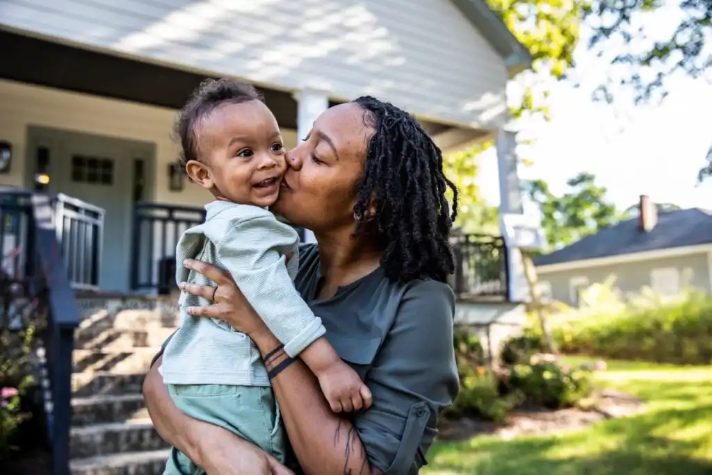 A woman stands outside a house, holding a smiling baby in her arms and kissing the baby on the cheek. The scene is bright and cheerful with greenery and another house in the background.