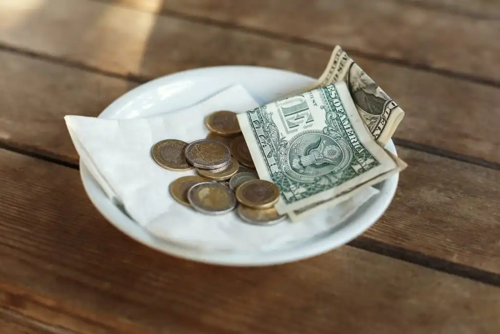 A white plate with coins and a folded one-dollar bill on a napkin, sitting on a wooden table, likely representing a tip at a restaurant.