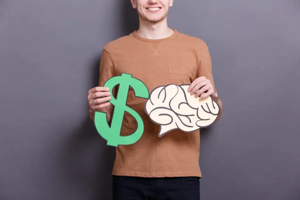A person in a brown long-sleeve shirt holds a large green dollar sign cutout in one hand and a paper cutout of a brain in the other, standing against a plain gray background.