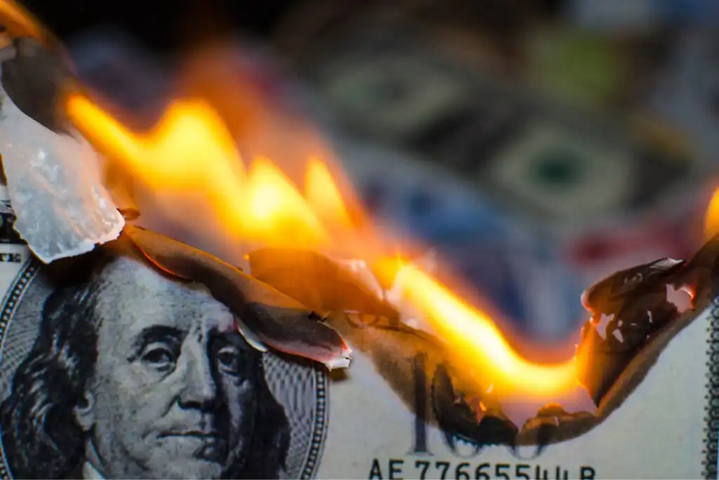 A close-up of a U.S. hundred-dollar bill burning, with bright flames consuming the paper and Benjamin Franklins face partially visible. Blurred currency can be seen in the background.