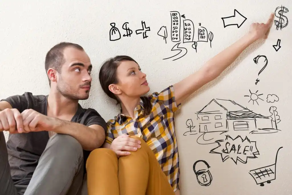 A man and woman sit on the floor, looking at hand-drawn illustrations of money, buildings, a house for sale, and question marks on the wall, symbolizing planning or dreaming about buying a home.