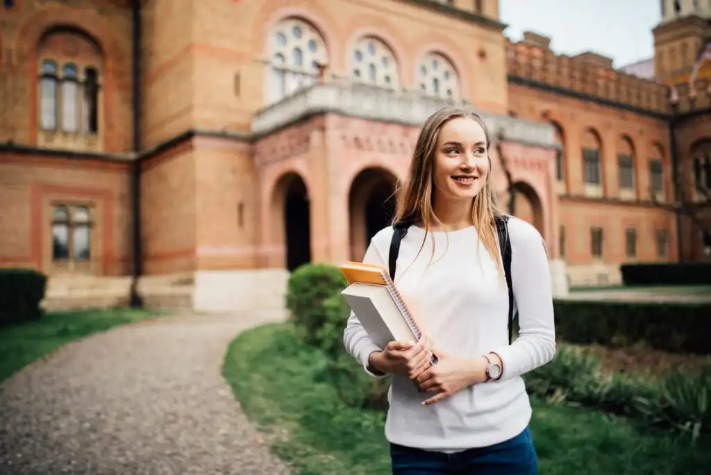 Young woman with long hair, wearing a white shirt and jeans, smiles while holding books and a notebook outside a large, historic brick building with arched windows and greenery.
