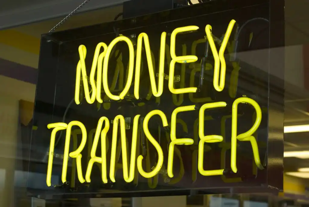 A glowing yellow neon sign reads Money Transfer, displayed in a window, indicating a place where people can send or receive money.