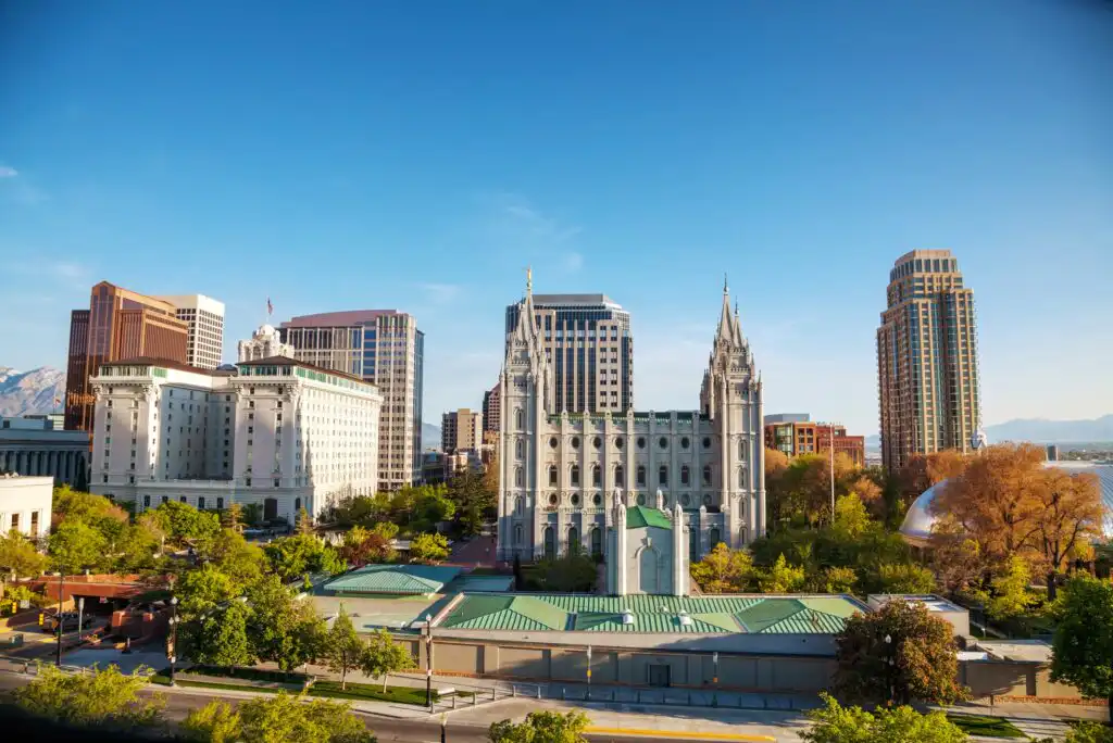 A cityscape featuring Salt Lake Citys downtown with the historic Salt Lake Temple at the center, surrounded by modern office buildings and lush green trees under a clear blue sky.