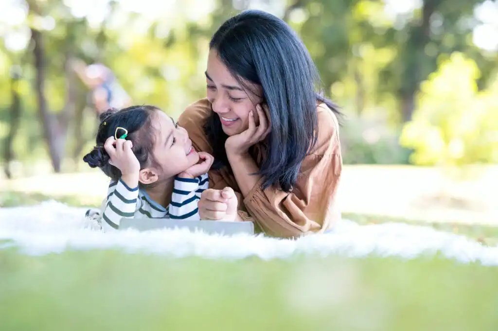 A woman and a young girl lie on a blanket outdoors, smiling and looking at each other. The scene appears bright and cheerful, with green grass and trees in the background.