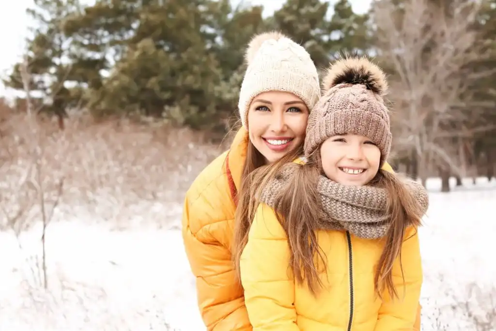 A smiling woman and a young girl, both wearing yellow winter jackets, knit hats, and scarves, stand close together outdoors in a snowy landscape with trees in the background.