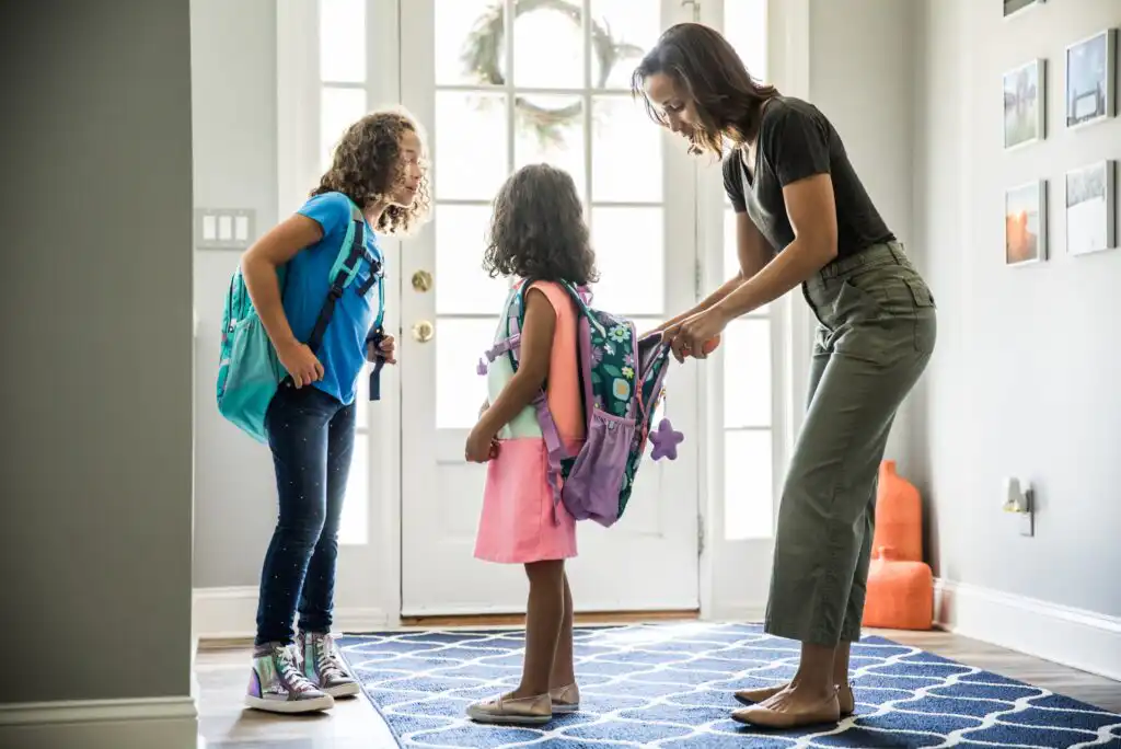 A woman helps a young girl with her backpack while another girl stands nearby, both children ready for school, in a bright entryway with a patterned blue rug.
