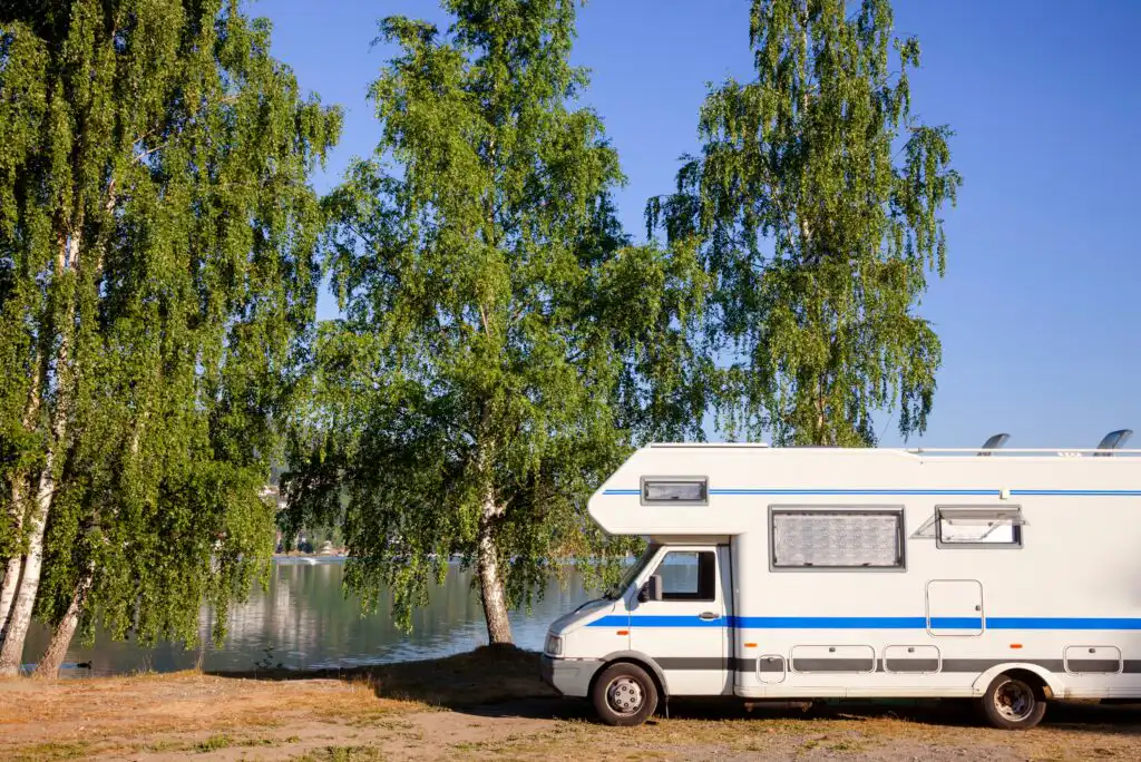 A white camper van is parked near a lake, surrounded by tall green trees on a sunny day with a clear blue sky.