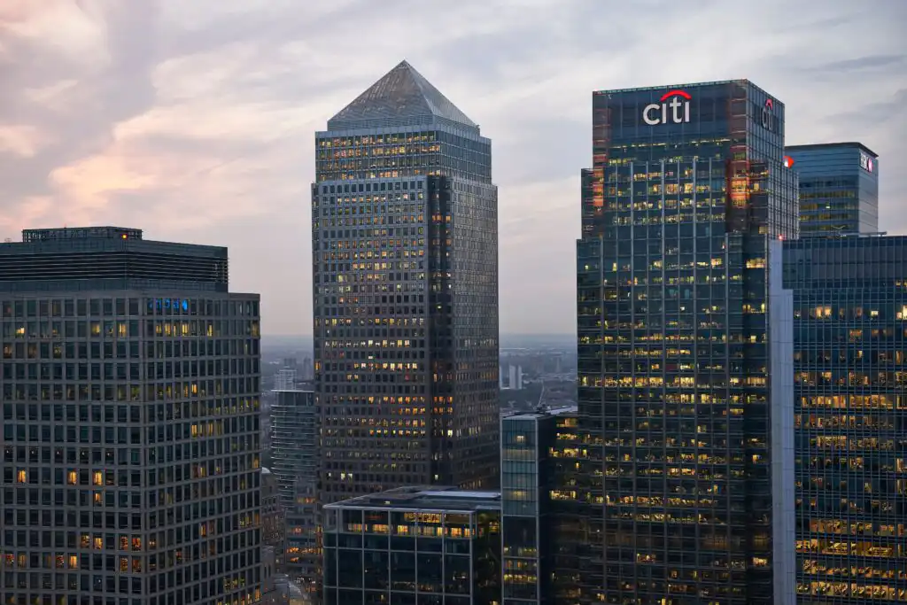 Skyscrapers in a city at dusk, including a glass building with the citi logo, all illuminated with office lights against a cloudy evening sky.