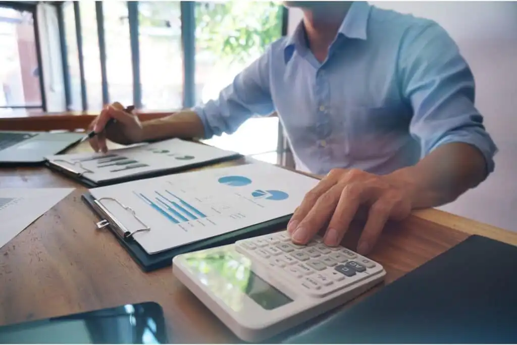 A person in a blue shirt sits at a desk reviewing financial charts on paper, using a calculator, with a smartphone and laptop nearby. Sunlight streams in through large windows in the background.