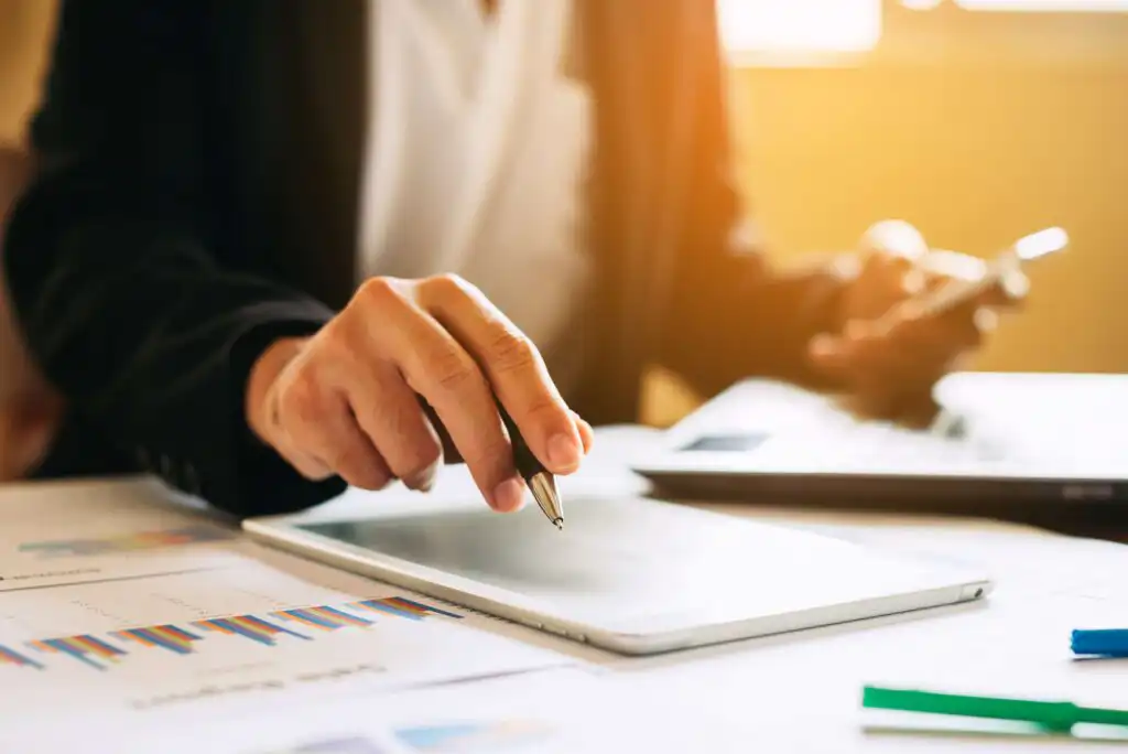 A person in business attire holds a pen over a tablet, with charts and graphs on paper nearby, and a laptop and smartphone in the background, suggesting a work or analysis setting.