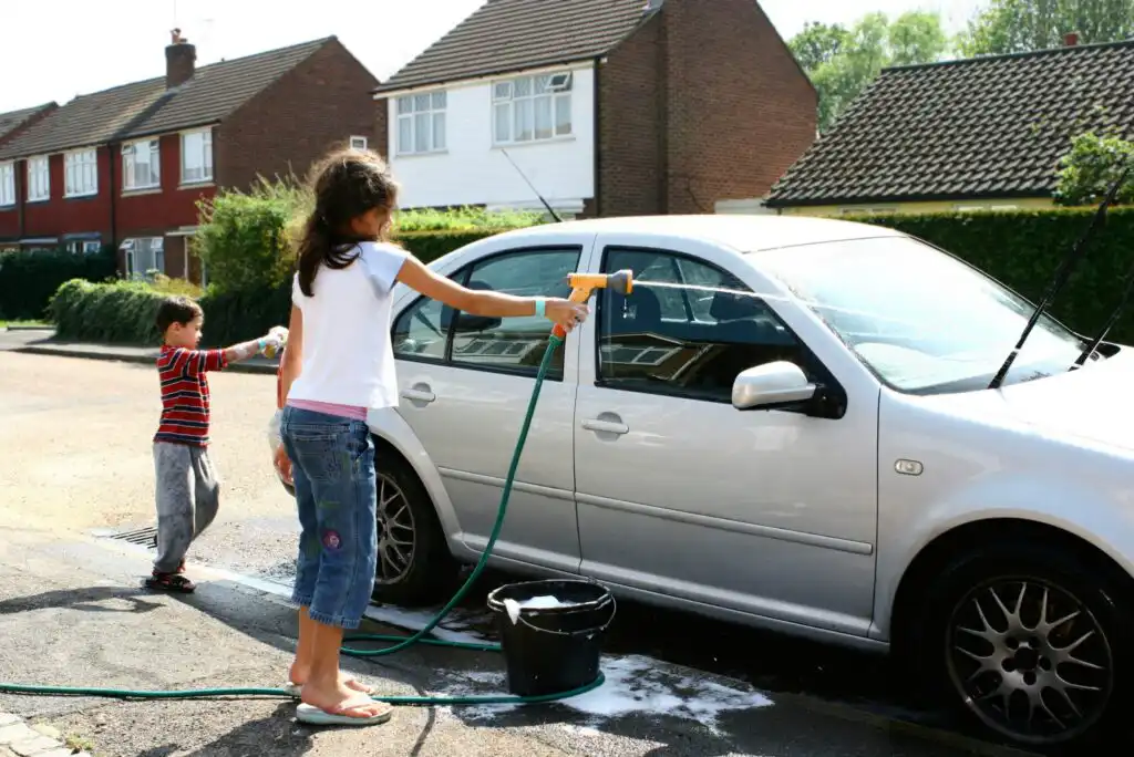 Two children are washing a silver car on a suburban street. The older child is using a hose while the younger sprays water. A black bucket with soapy water is on the ground next to them. Houses line the background.