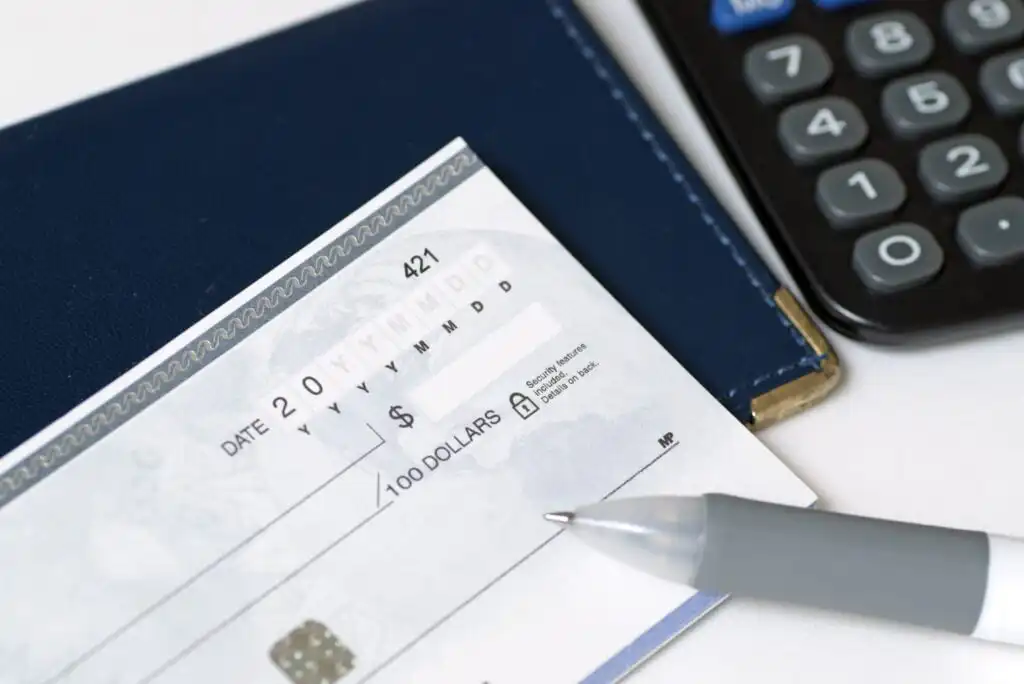 A close-up of a pen resting on a blank check with a blue checkbook holder and part of a calculator visible in the background.