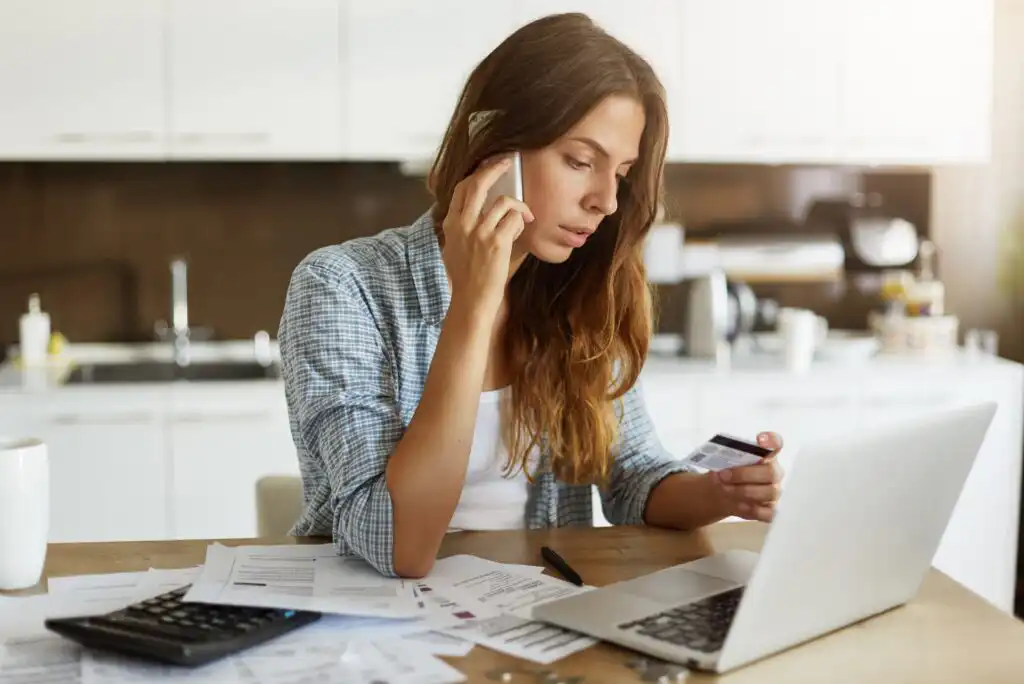 A woman sits at a table with papers and a calculator, talking on the phone while looking at a credit card and a laptop, appearing focused and concerned in a bright kitchen setting.
