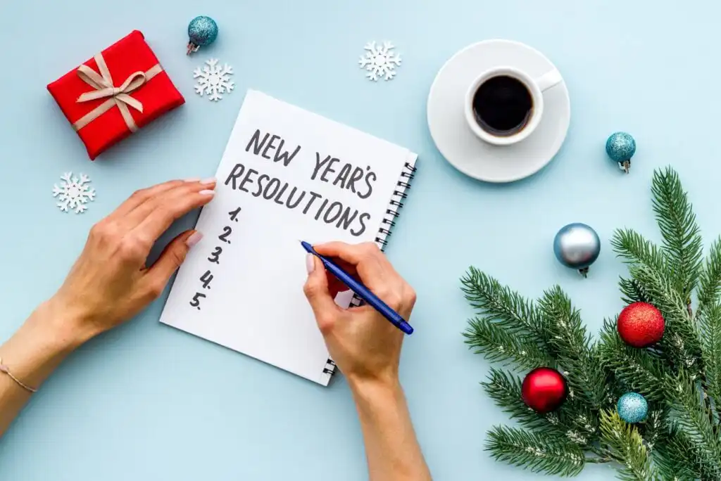 A person writes a list titled New Years Resolutions in a notebook on a blue table with a coffee cup, red gift box, Christmas ornaments, pine branches, and snowflake decorations.