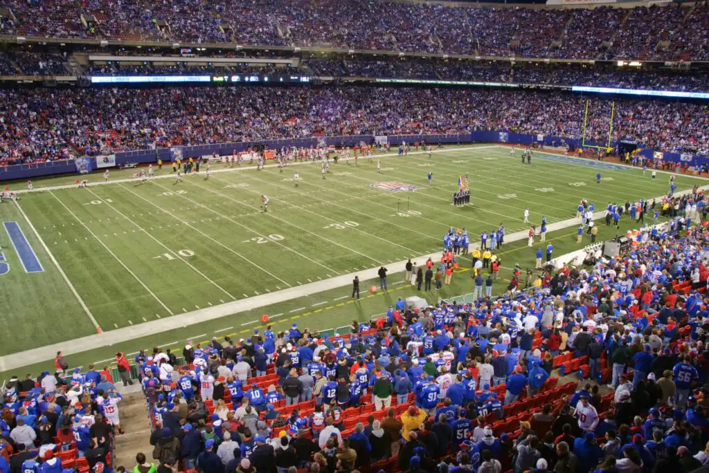 A crowded stadium with fans watching an American football game, players on the field, and coaches, photographers, and staff along the sidelines. The stands are filled with spectators wearing team colors.