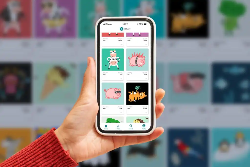 A hand holds a smartphone displaying a digital art marketplace with cartoon animal illustrations, including pigs and a tiger, on the screen. The person is wearing a red sweater. Blurred colorful images are in the background.