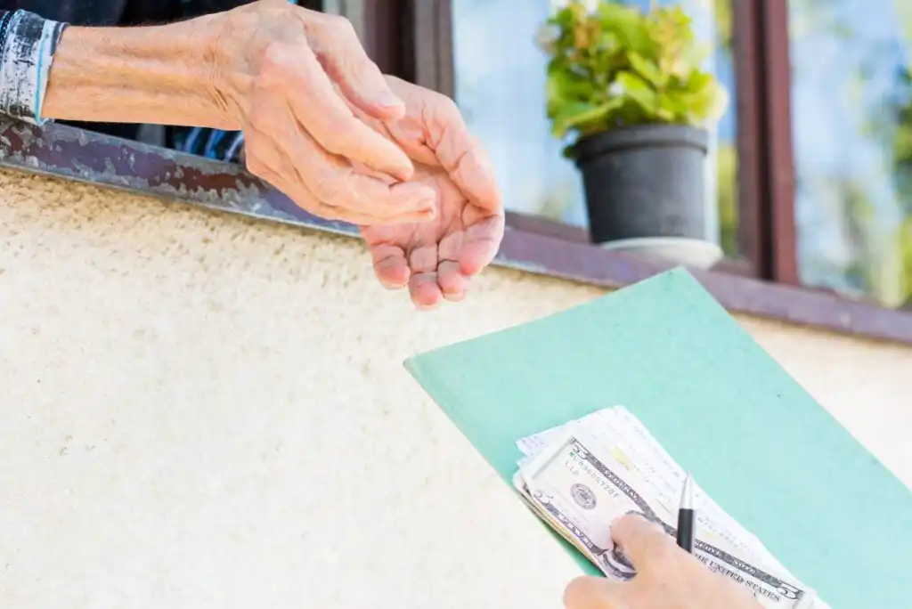 An elderly person reaches out to receive cash from another person holding money and a folder, with a potted plant and a window in the background.