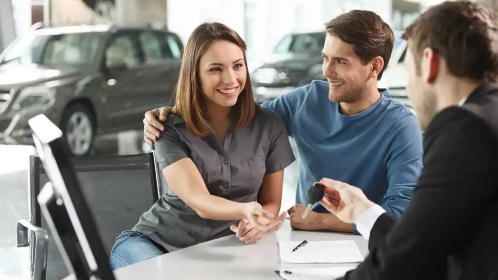 A smiling couple sits across a desk from a car dealer, who is handing them a set of car keys. Cars are visible in the background, suggesting they are at a dealership finalizing a purchase.