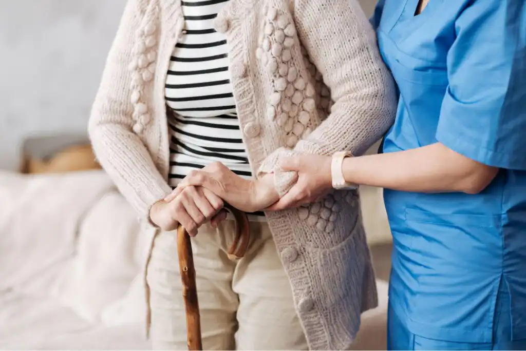 A caregiver in blue scrubs assists an elderly person wearing a striped shirt and knitted cardigan, who is holding a wooden cane. The caregiver gently supports the persons arm indoors.