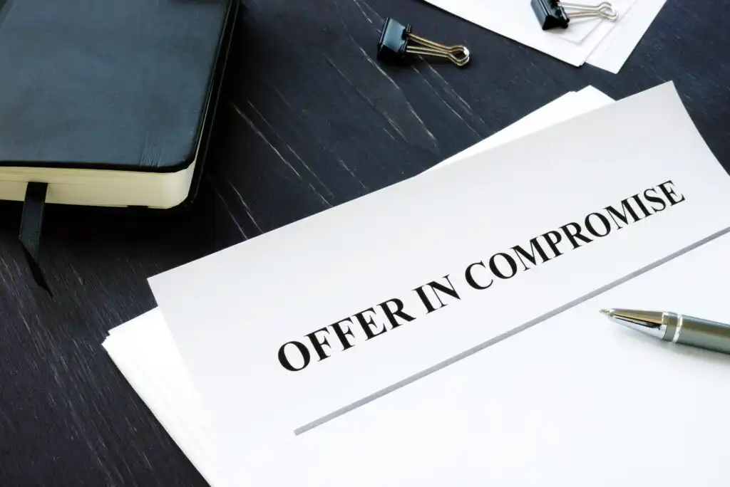 A document titled OFFER IN COMPROMISE sits on a dark desk with a pen nearby, next to a notebook, binder clips, and other papers.