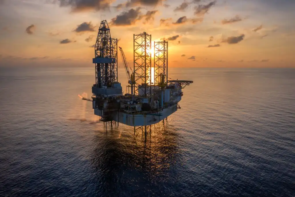 An offshore oil drilling platform stands in the ocean at sunset, with the sun partially hidden behind the structure and clouds scattered across the colorful sky.