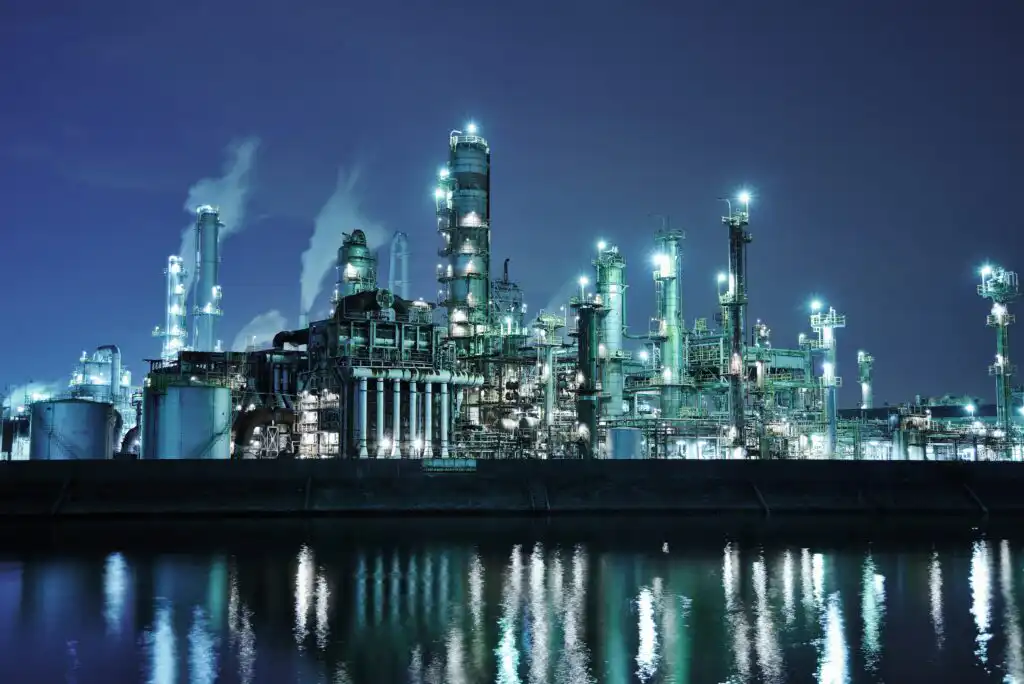 A brightly lit oil refinery or industrial plant at night, with smoke rising from stacks, reflected in the calm water in the foreground.