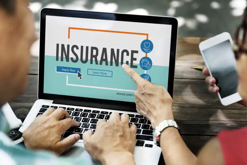 Two people sit at a table, one using a laptop displaying the word INSURANCE with buttons labeled Apply Now and Learn More, while the other holds a smartphone.