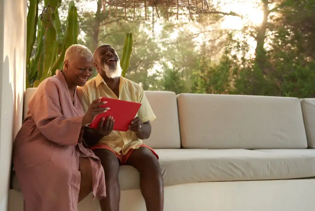 An older couple sits on an outdoor sofa, smiling and laughing together while looking at a red tablet, surrounded by greenery and sunlight filtering through the trees.