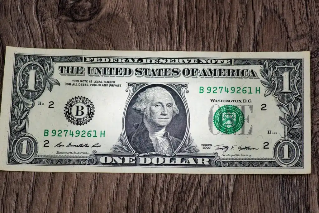 A United States one-dollar bill featuring George Washington’s portrait lies flat on a wooden surface. The bill displays serial numbers, treasury seal, and other standard design elements.
