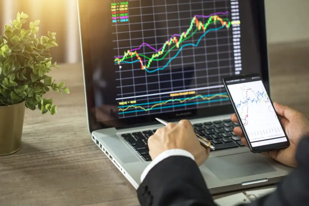 A person in a suit analyzes financial charts on a laptop and smartphone at a desk with a plant, focusing on stock market trends and data.
